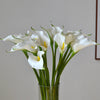 A bouquet of white artificial calla lily stems in a glass vase against a light background.