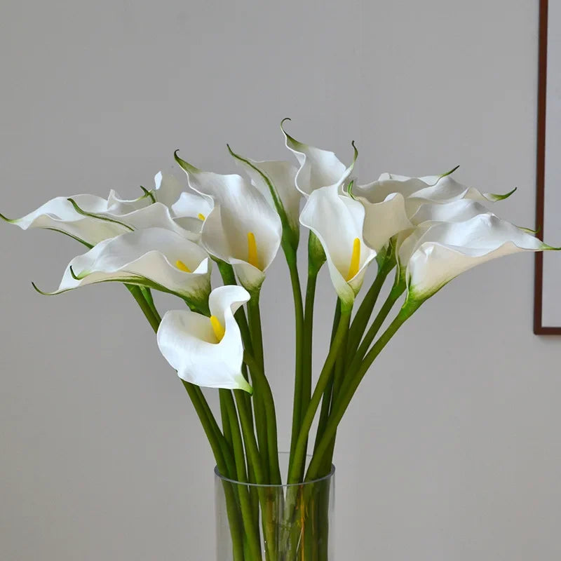 A bouquet of white artificial calla lily stems in a glass vase against a light background.
