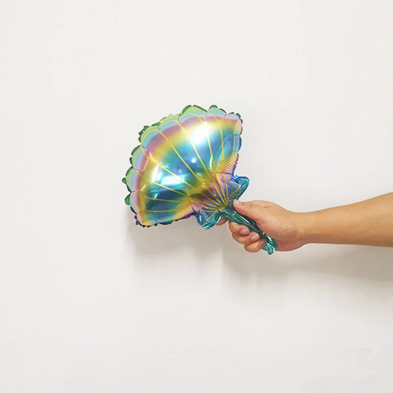 A hand holds a iridescent foil balloon shaped like a sea creature against a white wall.