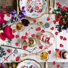 A rustic table setting featuring pink, red, and purple fabric heart petals scattered around a plate of sliced cake.