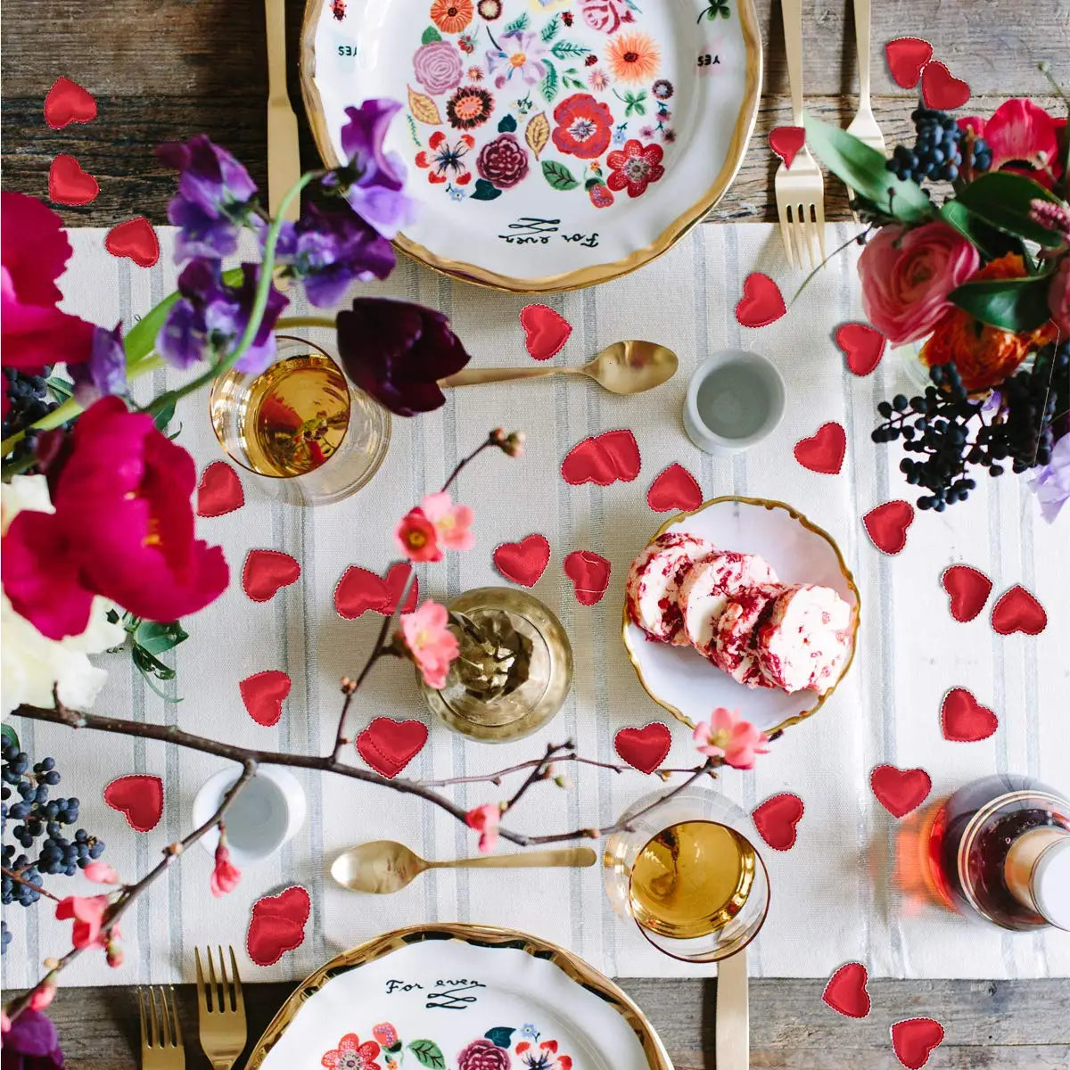 A rustic table setting featuring pink, red, and purple fabric heart petals scattered around a plate of sliced cake.