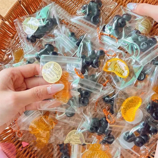A hand holds a collection of handmade fruit keychains in clear plastic bags on a woven tray.
