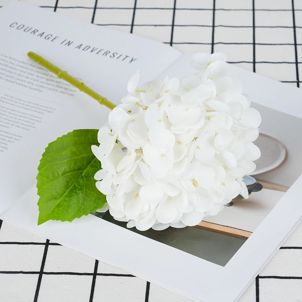 A close-up photo of a white artificial hydrangea flower cluster resting on a piece of paper with text.