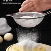 A hand holds a 3-piece set of stainless steel spoons and colanders, with powder falling onto a pastry on a wooden board.