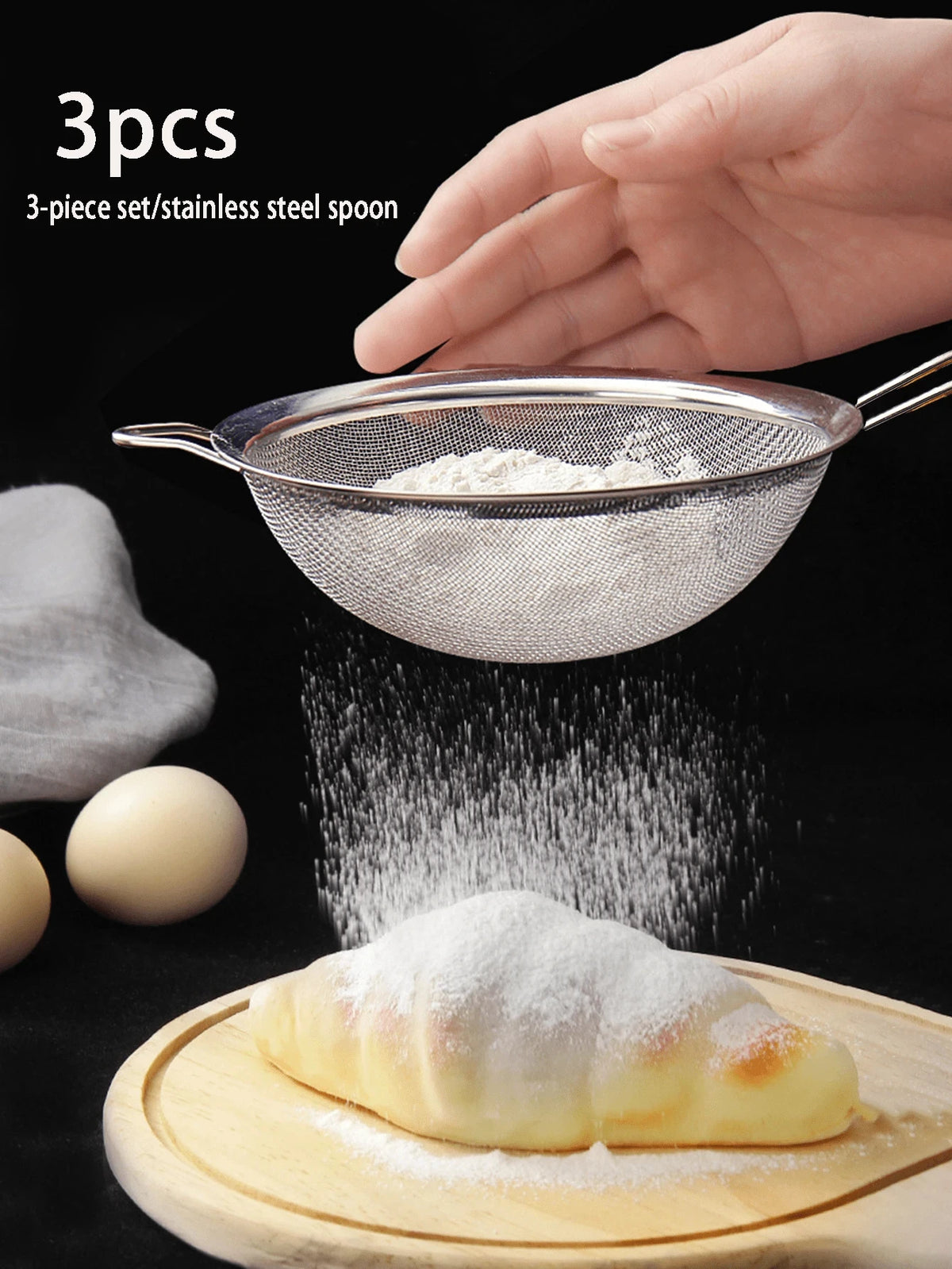 A hand holds a 3-piece set of stainless steel spoons and colanders, with powder falling onto a pastry on a wooden board.