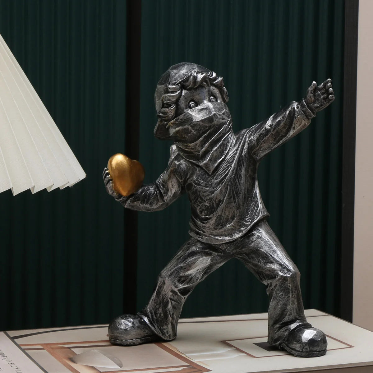 A gray resin sculpture of a child figure throwing a golden heart, set against a dark green background.