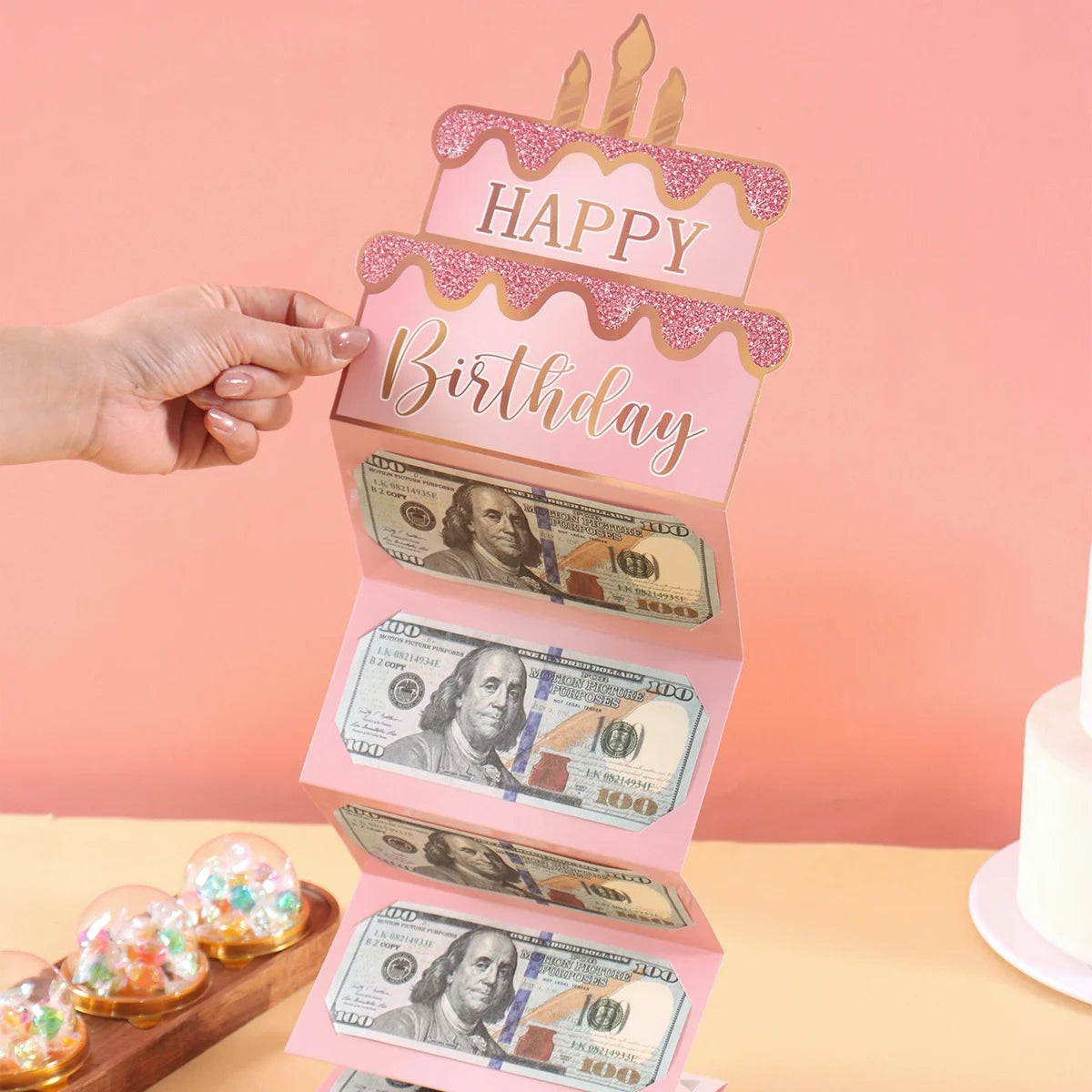 A tiered pink birthday cake design featuring cash gift envelopes and a topper.