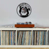 A vintage vinyl record wall clock with a black face and hands is mounted on a light gray wall above a wooden turntable.
