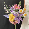 A handmade crochet flower bouquet featuring purple, pink, yellow, and white flowers arranged on a green stem.
