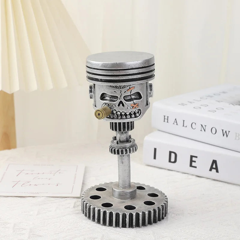 A silver mechanical piston skull sculpture with gears and a cylinder mounted on a circular base.