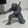 A gray miniature figure of a ninja in tactical gear holding a pen, displayed on a white surface.