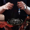 Hands holding a crystal pendulum over a wooden divination board with various symbols.