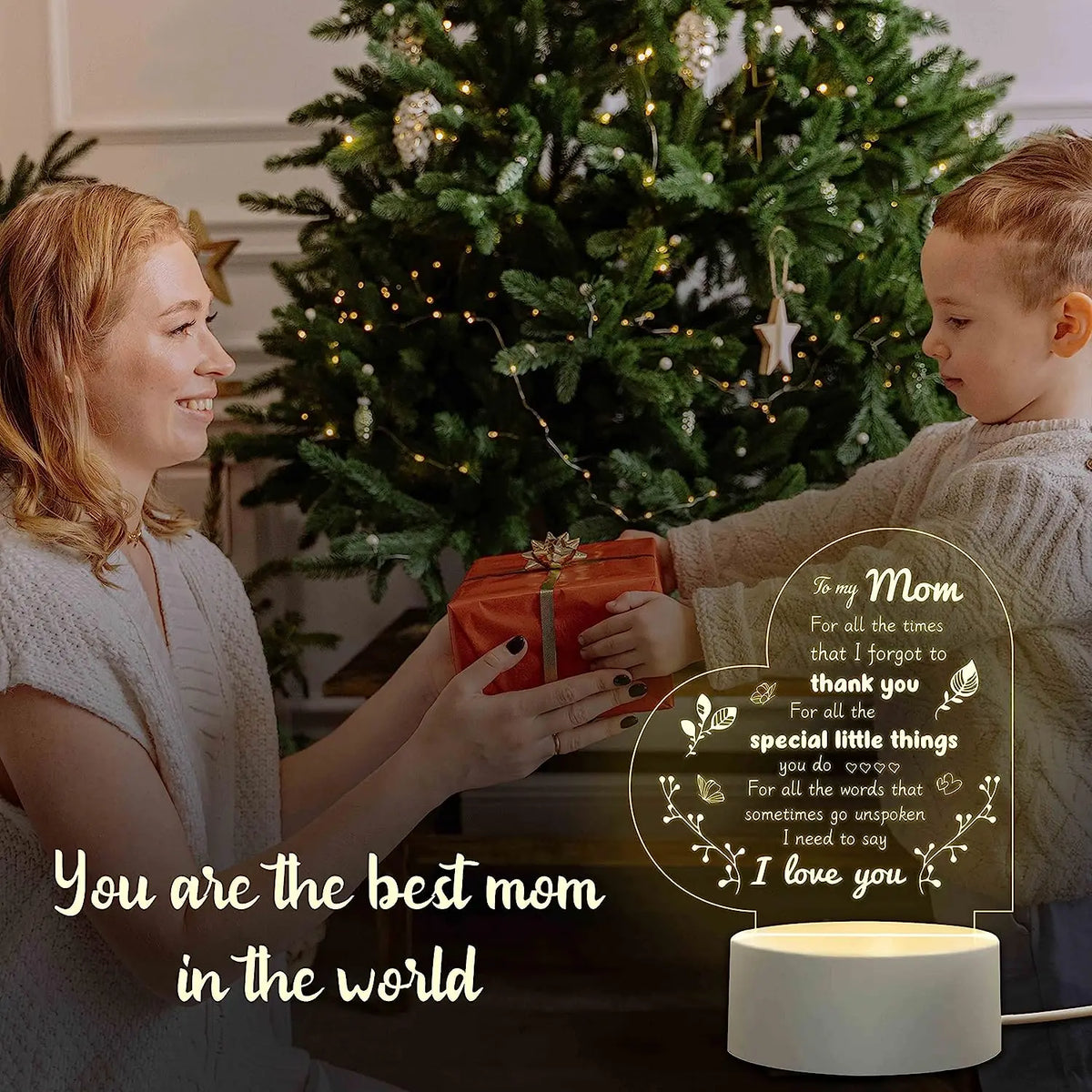 A woman gives a gift to her son in front of a decorated Christmas tree with lights.