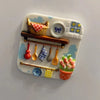 A white resin fridge magnet featuring a woven basket, various utensils, and a potted flower.
