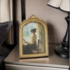 An ornate gold-framed vintage resin photo of a woman in period attire sitting and reading.