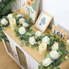 Künstlicher Eukalyptus- und Rosenkranz mit weißen Rosen und Kerzen auf einem Holzschranktopf.