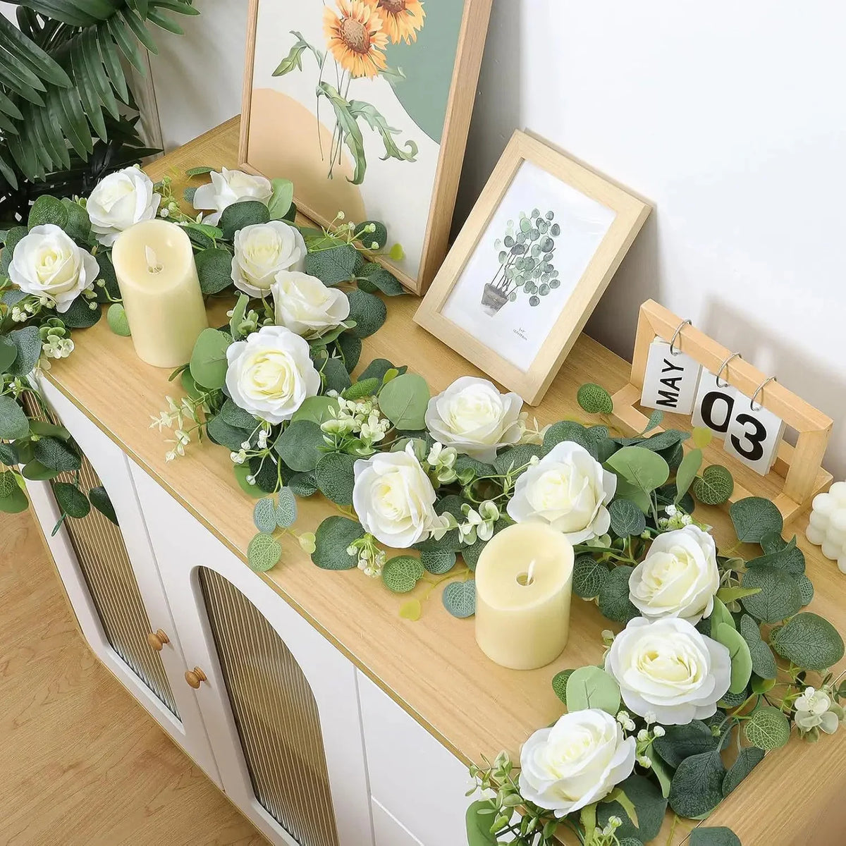 Künstlicher Eukalyptus- und Rosenkranz mit weißen Rosen und Kerzen auf einem Holzschranktopf.