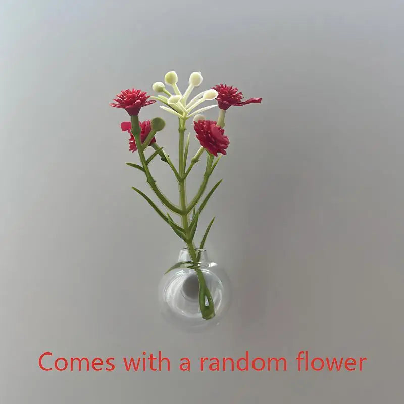 A small glass vase holds a plant with pink and white flowers on a light gray background.