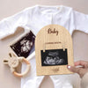 A wooden pregnancy announcement photo frame with the word Baby and Coming Soon on it, held against a white shirt.