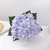 A cluster of light purple artificial hydrangea flowers resting on green leaves on a white surface.