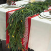 A long green Norfolk pine garland with small lights draped across a red table runner on a white tablecloth.