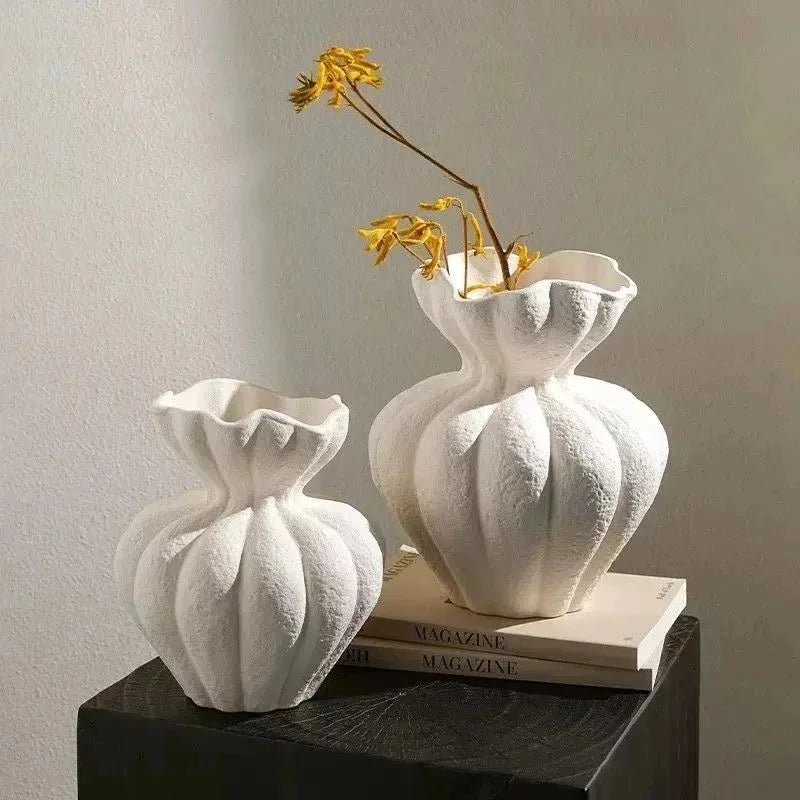 A white ceramic vase with a textured surface holds a sprig of dried yellow flowers on a dark wooden surface.