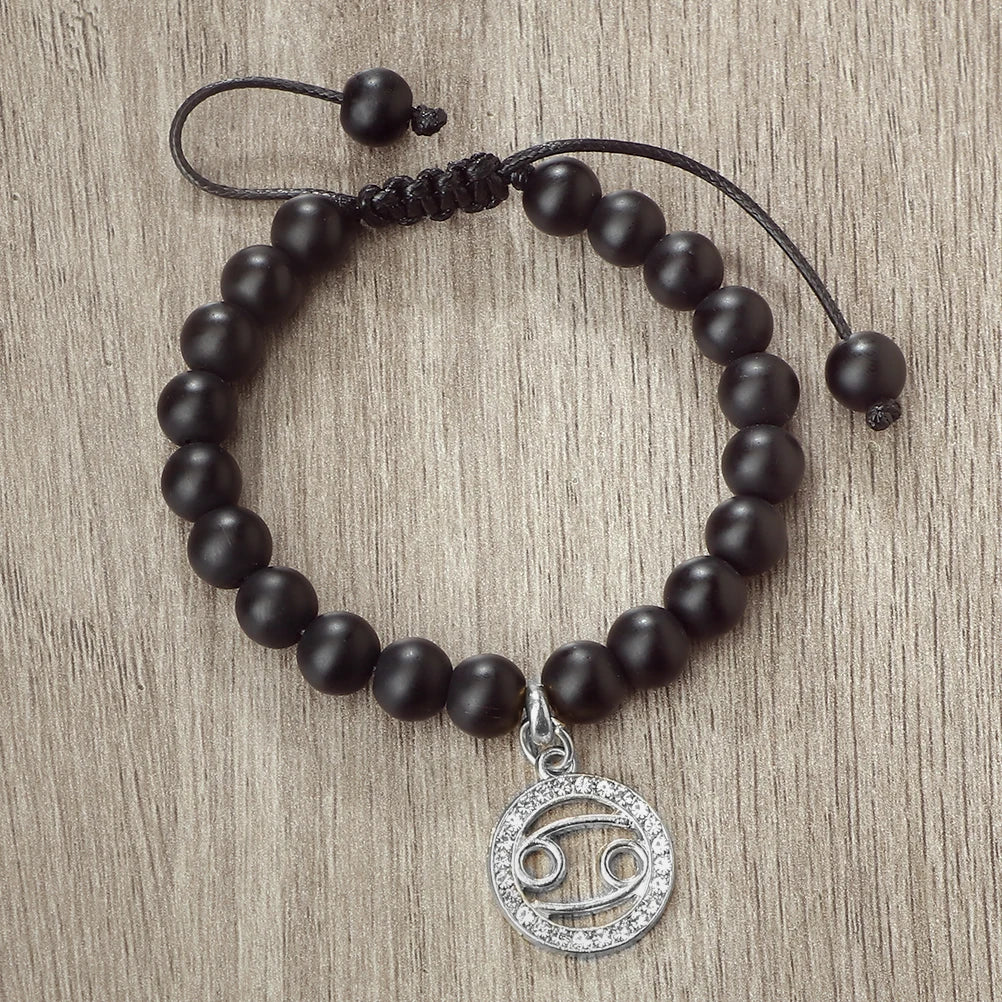 A black beaded bracelet with a silver zodiac sign charm is displayed on a wooden surface.