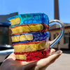 A stack of hand-painted book designs in blue, yellow, purple, and red on a clear glass mug being held in a hand.