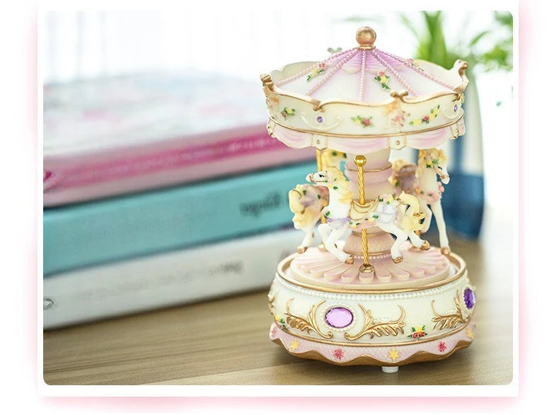 A delicate pink and cream handmade resin carousel music box with gold accents sits on a wooden surface.