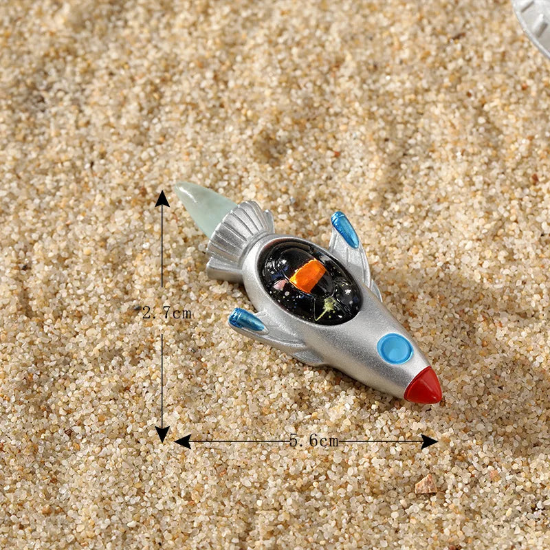 Décoration de gâteau d'astronaute miniature argentée avec des accents bleus repose sur du sable grossier.