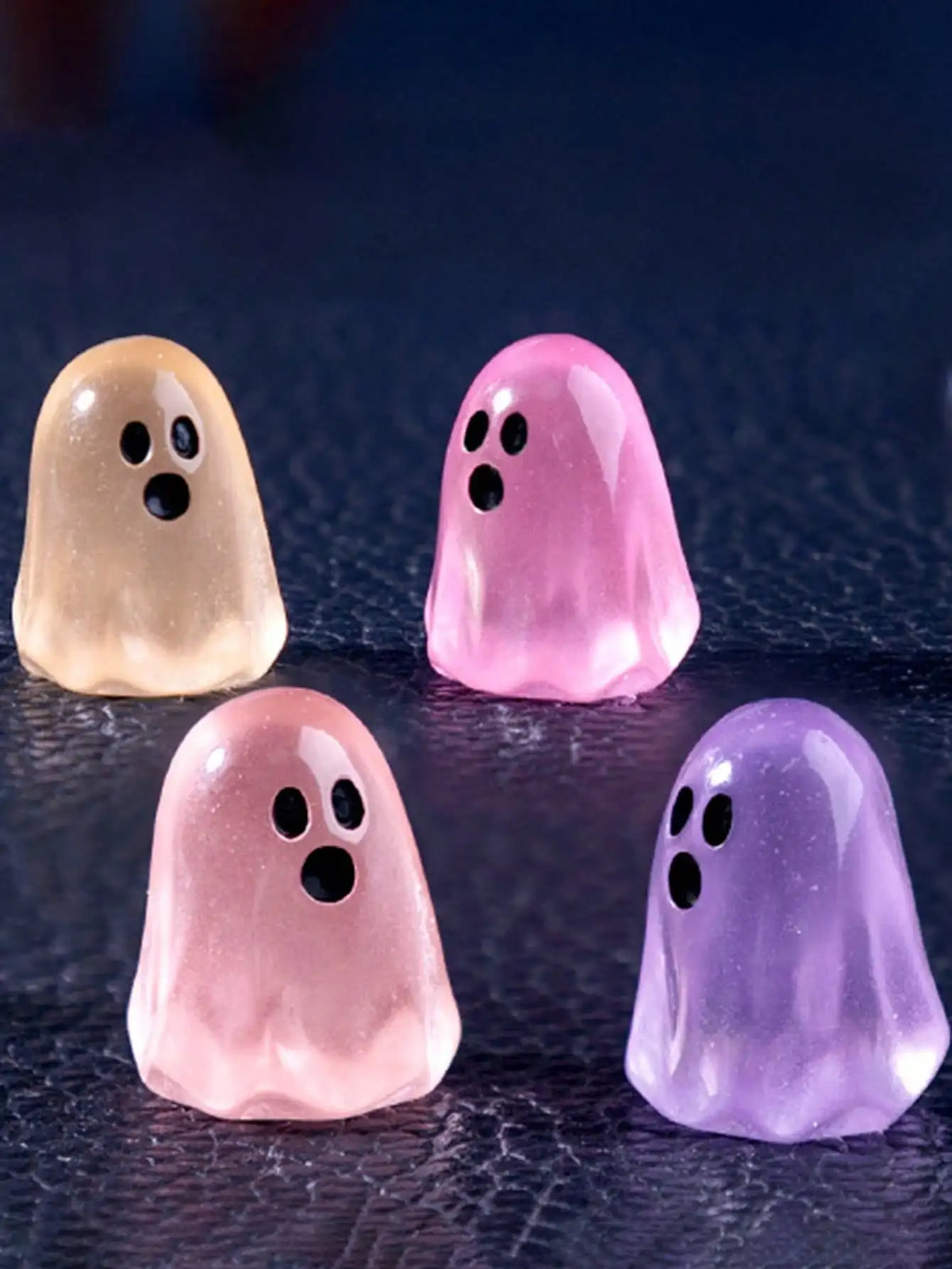 Four luminous ghost mini figurines in pastel shades of yellow, pink, and purple on a dark surface.