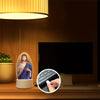 An illuminated Jesus Christ night light is displayed on a wooden surface next to a television screen.