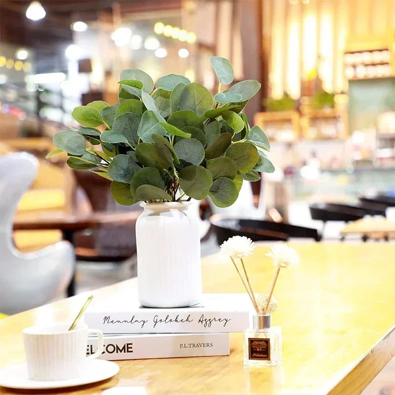A potted artificial eucalyptus plant in a white vase displayed on a wooden table.