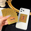 A hand holds gold wealth amulets and stickers next to a white smartphone.