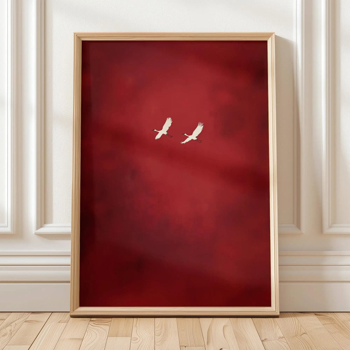 A framed canvas wall art featuring a deep red background with two white cranes, displayed on a light wooden floor.