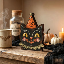 A carved wooden cat sculpture with orange and black paint sits on a rustic wooden surface next to a glass jar.