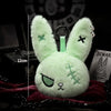 A light green bunny plush keychain with embroidered details resting on a dark red fabric.