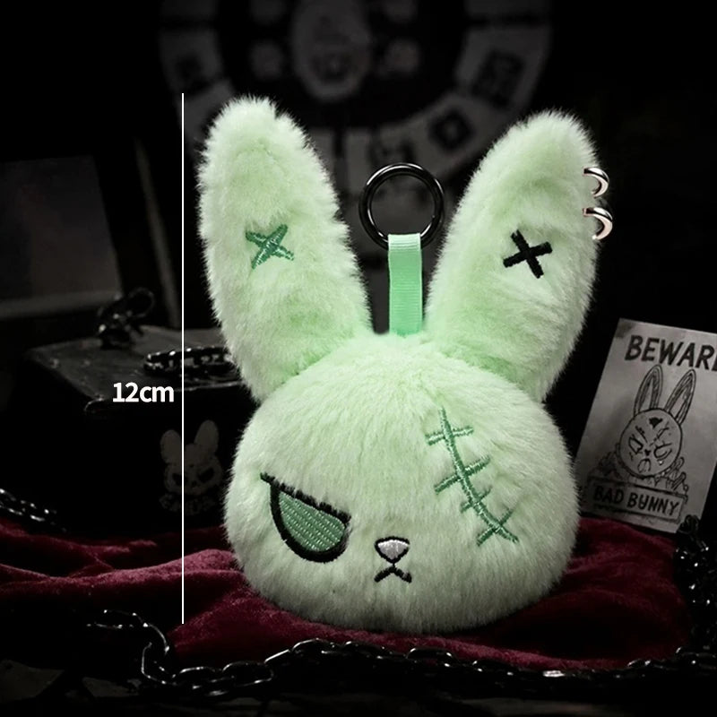 A light green bunny plush keychain with embroidered details resting on a dark red fabric.