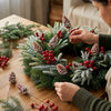 Hands arrange artificial Norfolk pine picks with red berries and pine cones on a wooden surface.