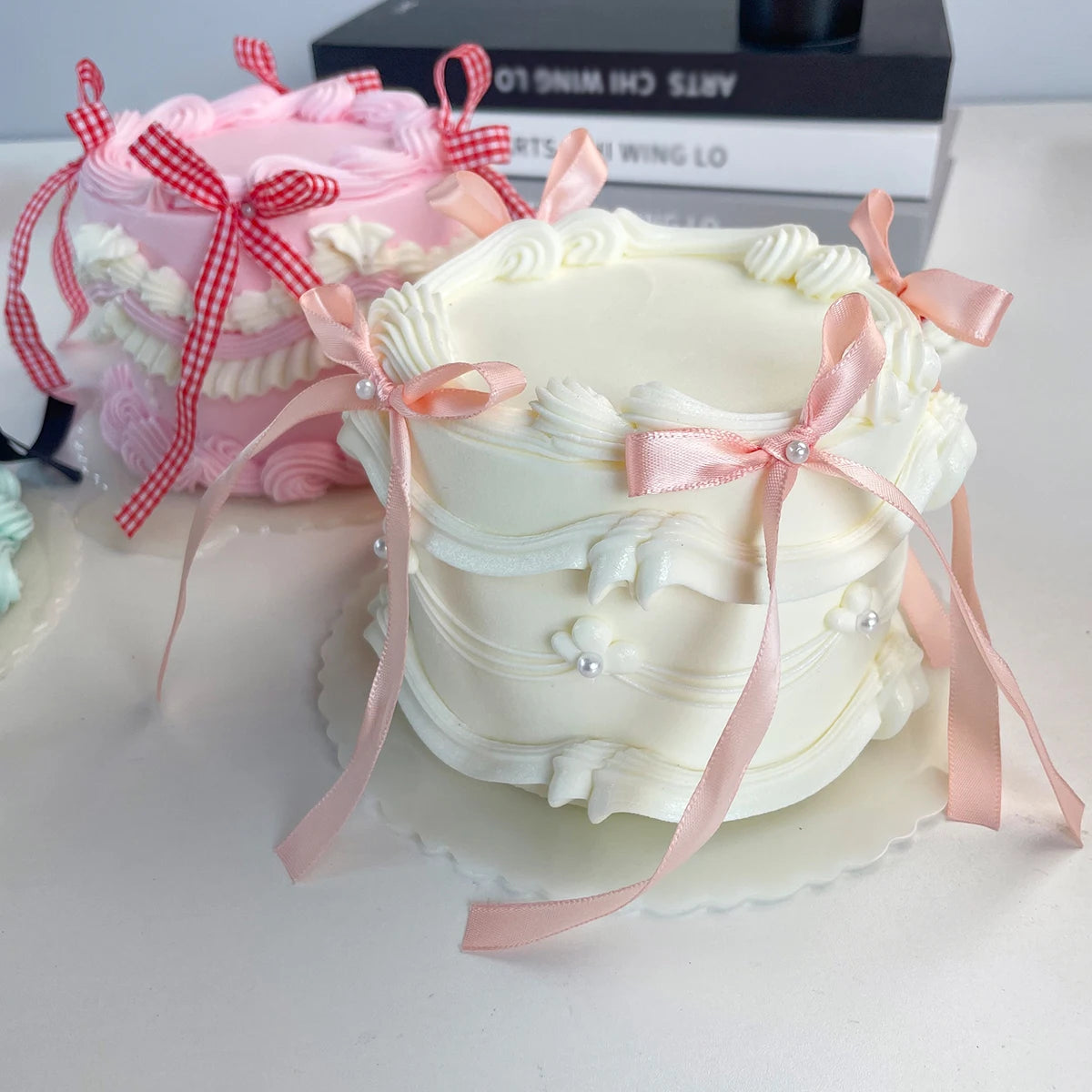 A white decorative fake cake with pink ribbons and flowers is displayed next to a pink cake.