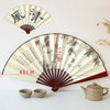 A large, folding decorative fan with a bamboo and silk design is displayed next to a traditional teapot.