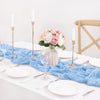 Light blue cheesecloth table runner with white plates, silverware, and tall candles set for a rustic event.