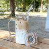 A tall glass mug filled with iced coffee or milk tea is resting on a wooden surface next to its clear lid and straw.