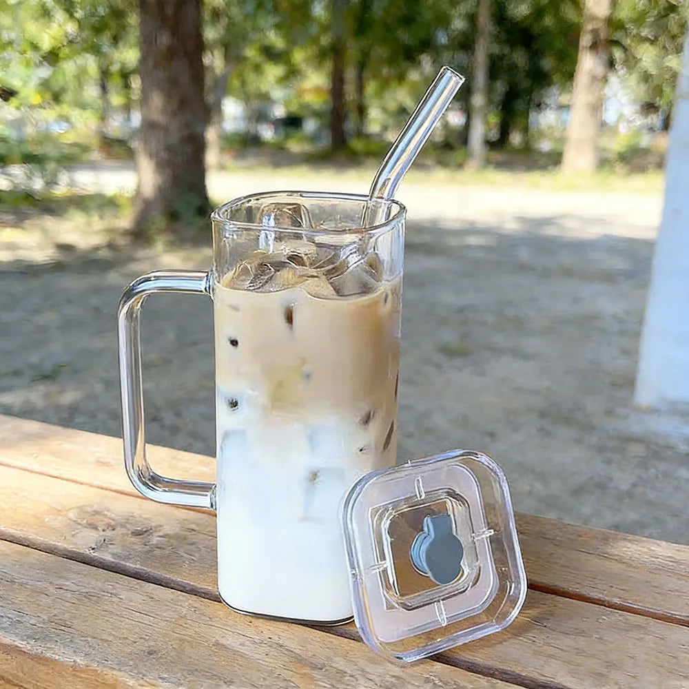 A tall glass mug filled with iced coffee or milk tea is resting on a wooden surface next to its clear lid and straw.