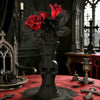 A dark gothic decorative vase shaped like a skull holding red roses on a red table.