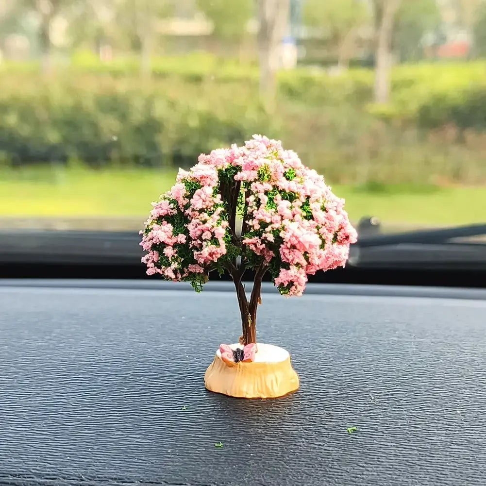 A small tree ornament with pink flowers sits on a black car dashboard.