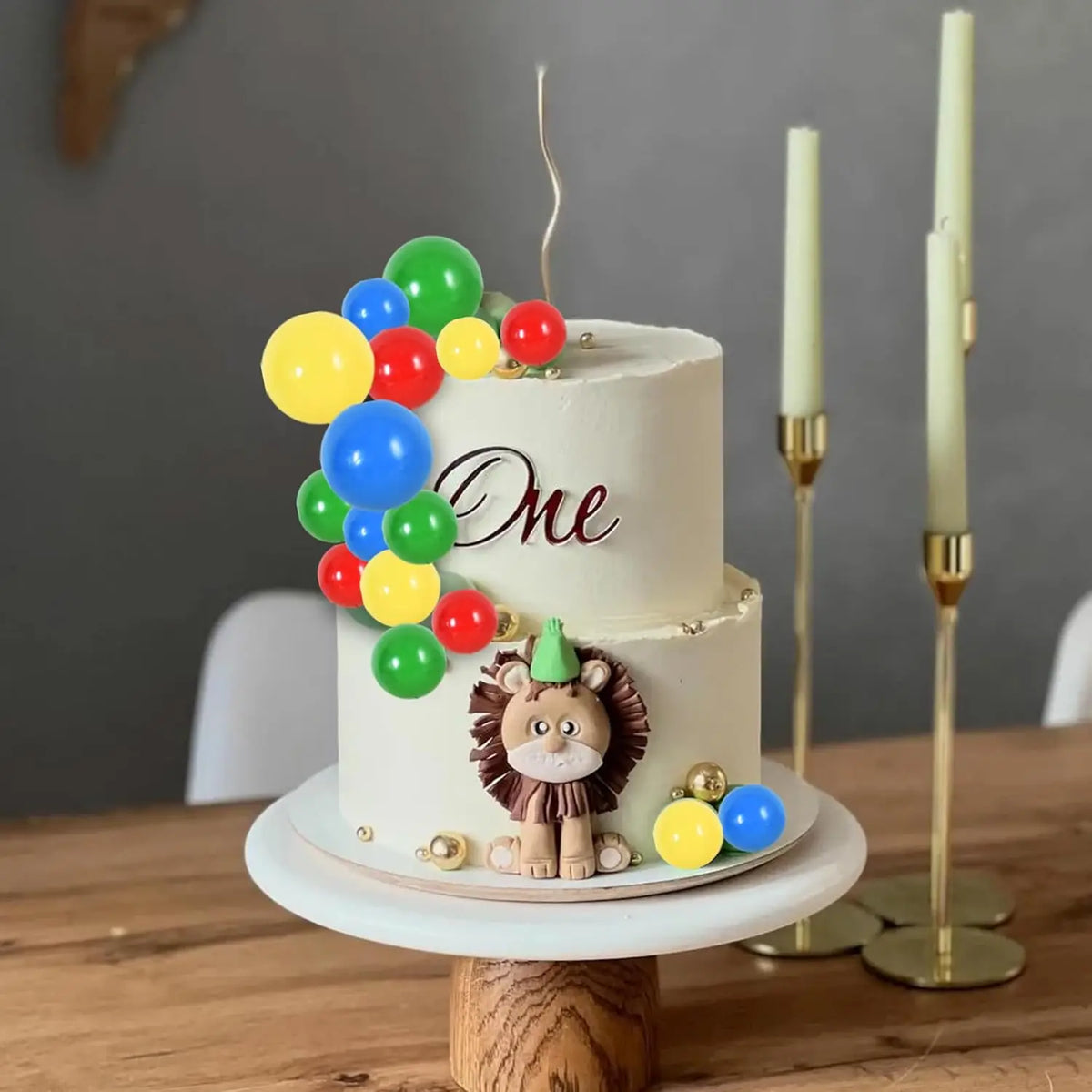 A two-tiered white cake decorated with colorful foam ball toppers and a lion figurine, displayed on a wooden stand.