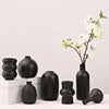 A collection of matte black ceramic vases and a branch with white flowers on a white surface.