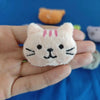 A small pink cat plush doll with black whiskers and pink stripes is held in a hand against a blue background.