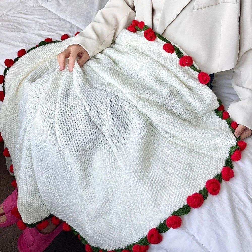 A white knitted blanket with a green trim and red floral accents is being held on a white bed.